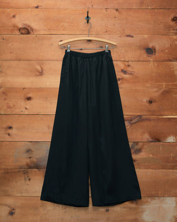 Roy Pants - Black, featuring a wide-leg silhouette, displayed on a wooden hanger against a rustic wooden wall background.