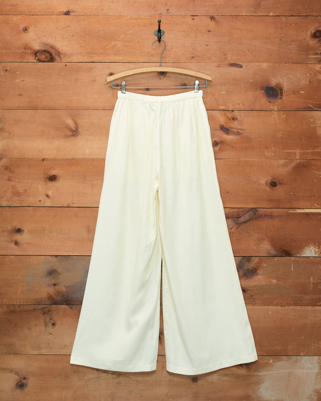 Roy Pants - White, wide-leg women’s trousers inspired by 1920s beach pajamas, hanging on a wooden hanger against a rustic wooden wall background.