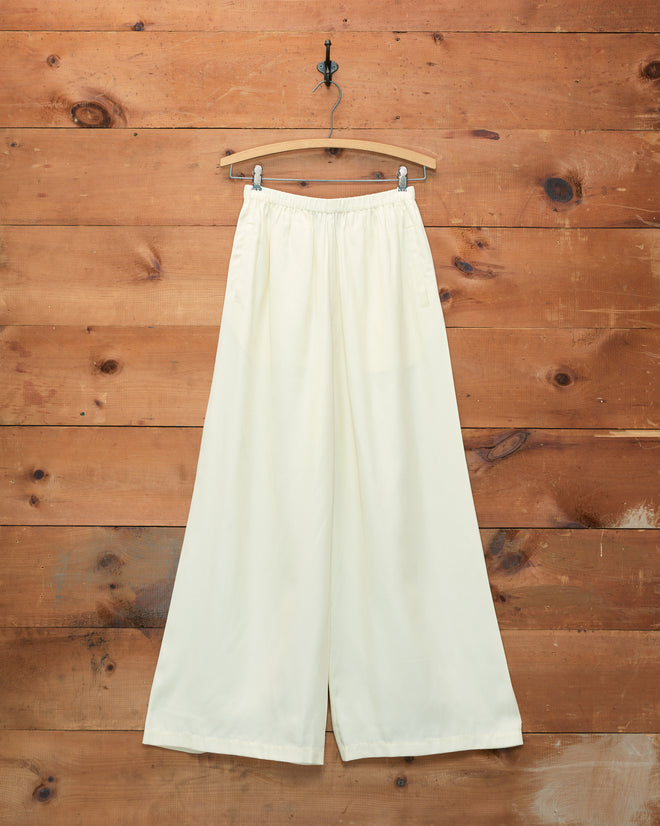 Roy Pants - White, inspired by classic 1920s beach pajamas, hang gracefully on a wooden hanger against a rustic wooden wall background.