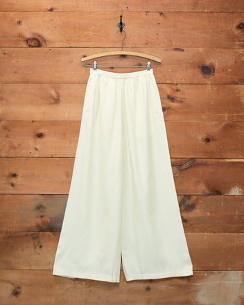 Roy Pants - White, inspired by classic 1920s beach pajamas, hang gracefully on a wooden hanger against a rustic wooden wall background.