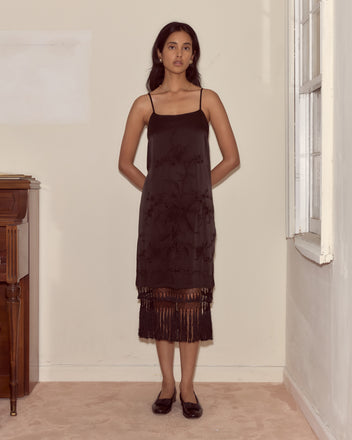 A woman in the Worcester Fringe Dress—embroidered silk with a crocheted fringe—stands in a beige room near a window and wooden cabinet.