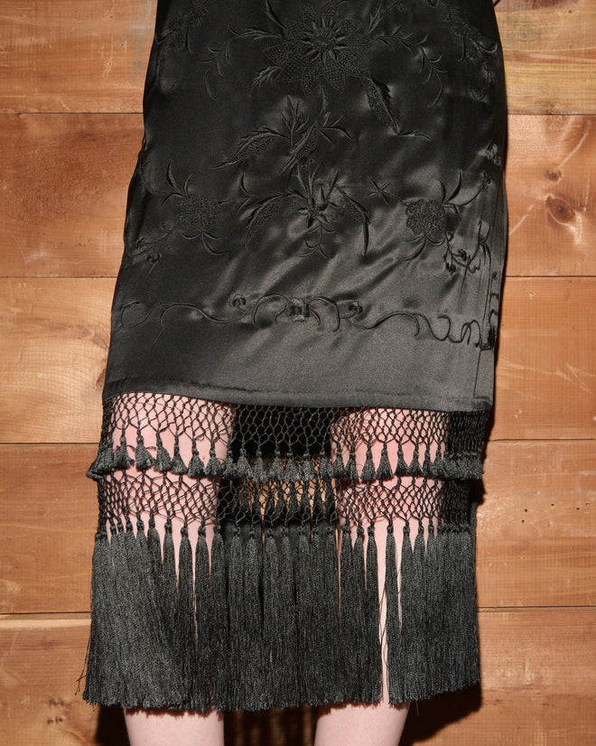 Worcester Fringe Dress in black, featuring embroidery and a net hem with fringe detail inspired by classic piano shawls, photographed in front of a wooden wall.