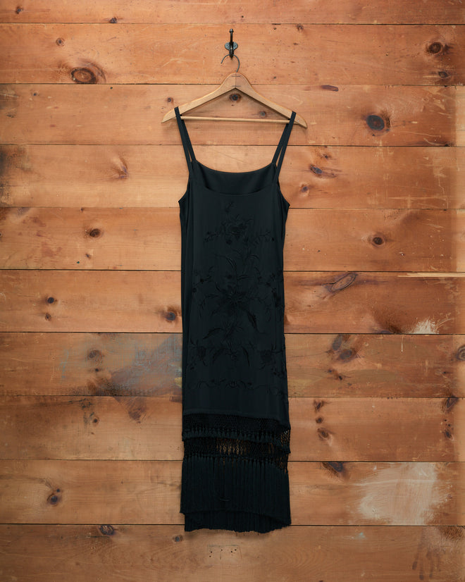 The Worcester Fringe Dress, a sleeveless black silk piece, hangs on a wooden hanger against a wooden plank wall.