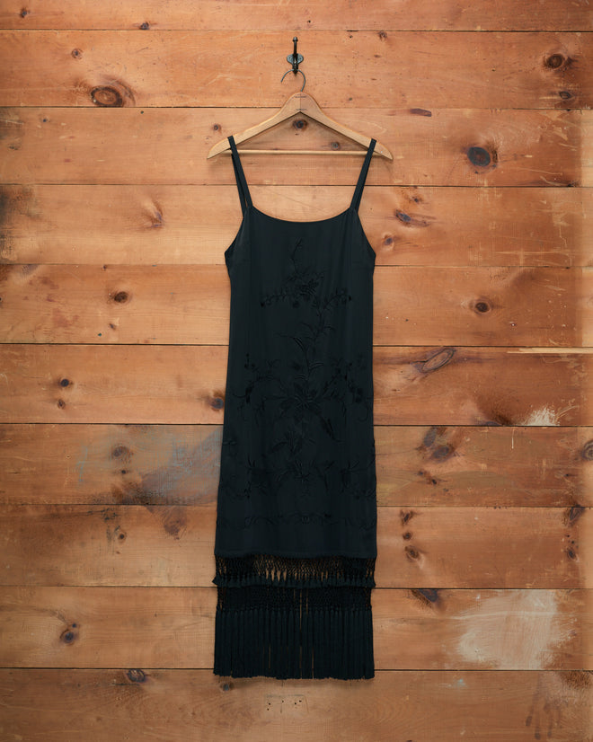 The Worcester Fringe Dress, a black sleeveless silk piece with a fringe hem, hangs on a wooden hanger against a wooden wall, evoking the elegance of a piano shawl.