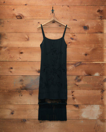 The Worcester Fringe Dress, a black sleeveless silk piece with a fringe hem, hangs on a wooden hanger against a wooden wall, evoking the elegance of a piano shawl.
