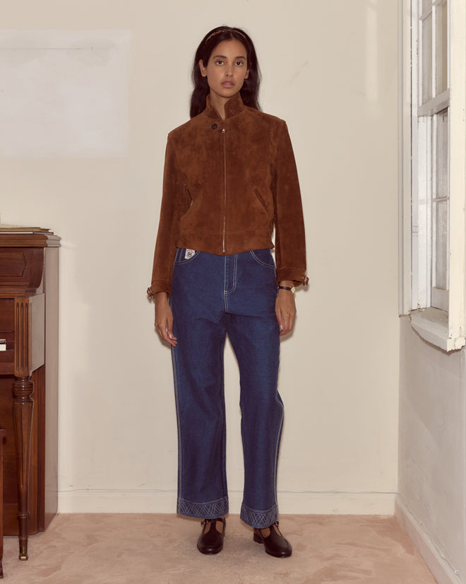 Woman stands indoors by a window, wearing the Suede Howard Jacket - Dark Brown, paired with blue jeans and black shoes.