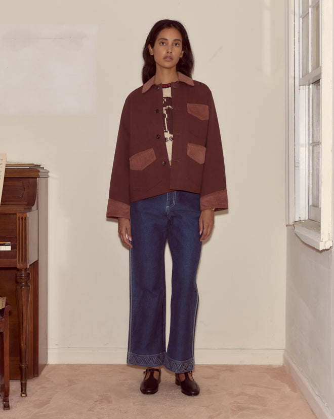 A woman stands indoors beside a piano and window, wearing the Craft Jacket - Brown with corduroy trim, blue jeans, and black shoes.