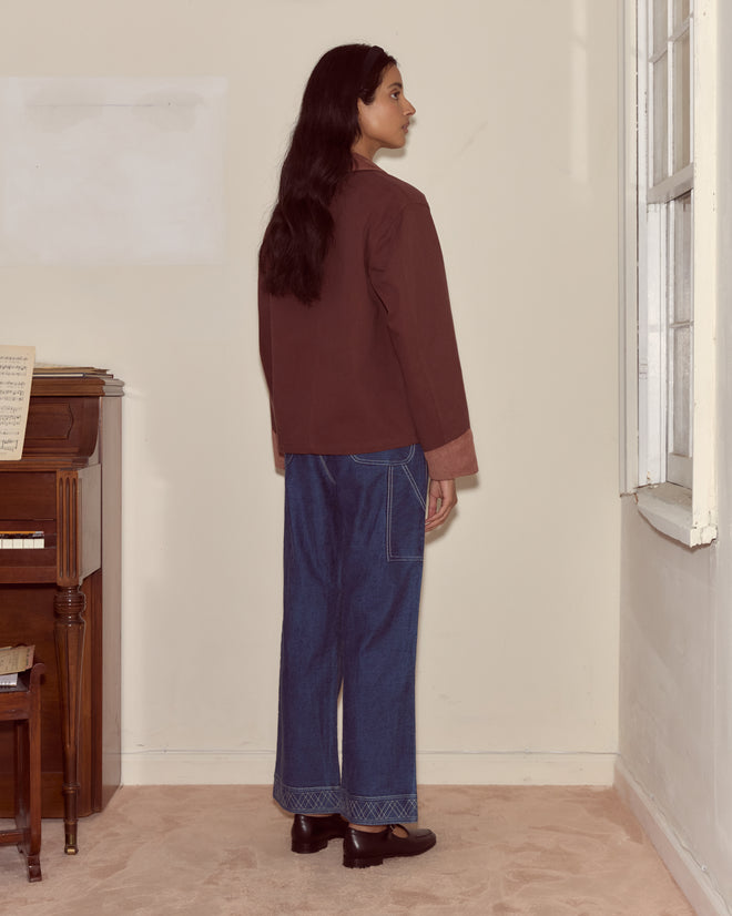 A person stands indoors facing sideways, wearing the Craft Jacket - Brown with corduroy trim, wide-leg jeans, and black shoes.