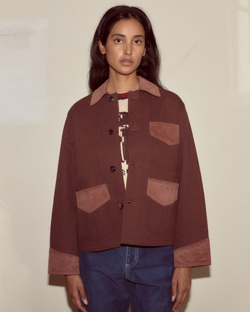 Woman wearing the Craft Jacket - Brown with large pockets and blue jeans, standing against a plain beige wall.