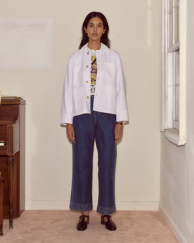 A woman stands indoors wearing the Craft Jacket - White, featuring cotton twill with corduroy trim, paired with a graphic tee, blue jeans, and black shoes.
