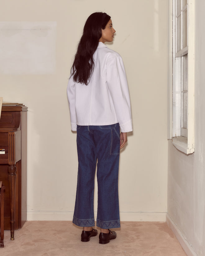A woman with long dark hair stands indoors facing a wall, wearing a white shirt, blue jeans, and the Craft Jacket - White.