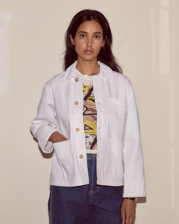 A woman wears the Craft Jacket - White, a cotton twill workwear style with corduroy trim, paired with jeans. She stands against a plain beige wall, looking directly at the camera.