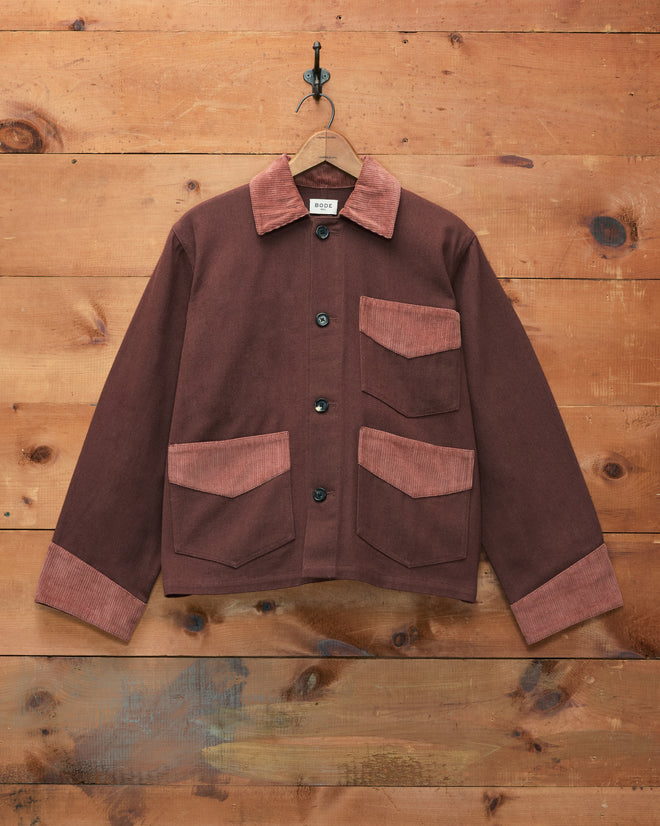 The Craft Jacket - Brown, made of cotton twill with lighter corduroy trim on the collar, cuffs, and pockets, hangs on a hook against a wooden wall.