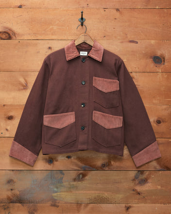 The Craft Jacket - Brown, made of cotton twill with lighter corduroy trim on the collar, cuffs, and pockets, hangs on a hook against a wooden wall.