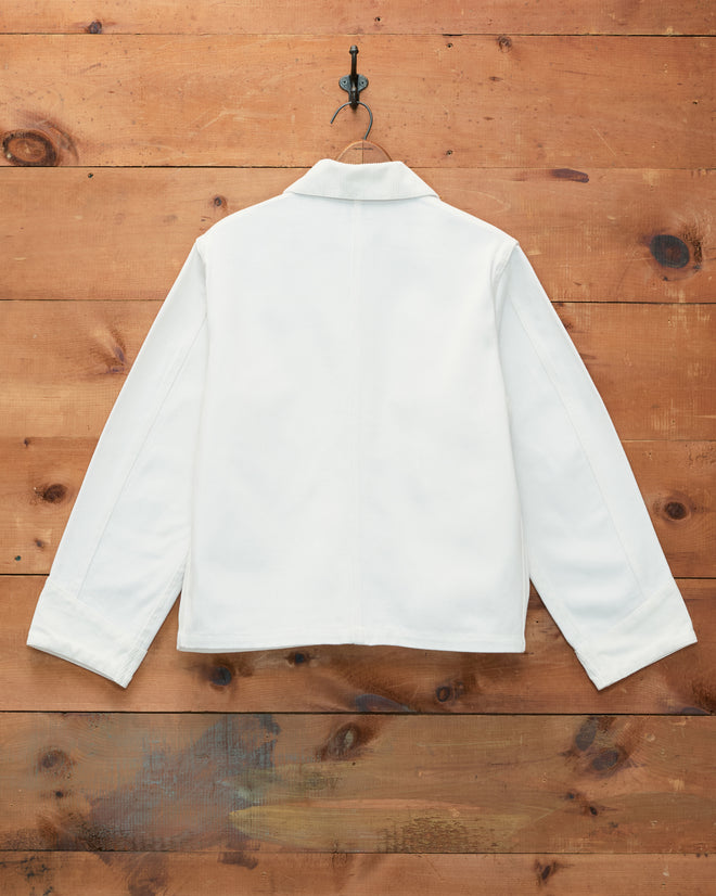 The Craft Jacket - White, featuring a boxy silhouette in cotton twill, is displayed from the back as it hangs on a black hook against a rustic wooden wall.