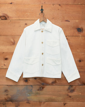 The Craft Jacket - White, a white cotton twill workwear jacket featuring a boxy silhouette and three front pockets, hangs on a hook against a wooden wall background.