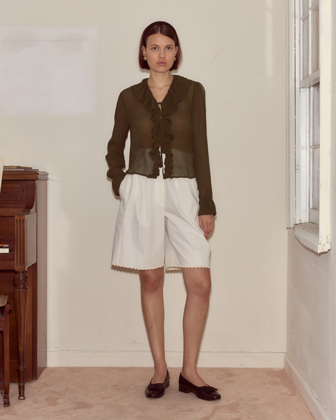 A woman stands indoors near a window and piano, wearing the Flounce Blouse - Olive and white shorts.