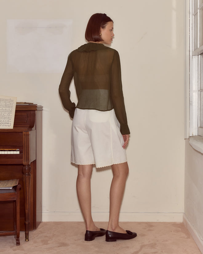 A woman stands indoors near a piano, facing away, wearing the Flounce Blouse - Olive and white shorts.
