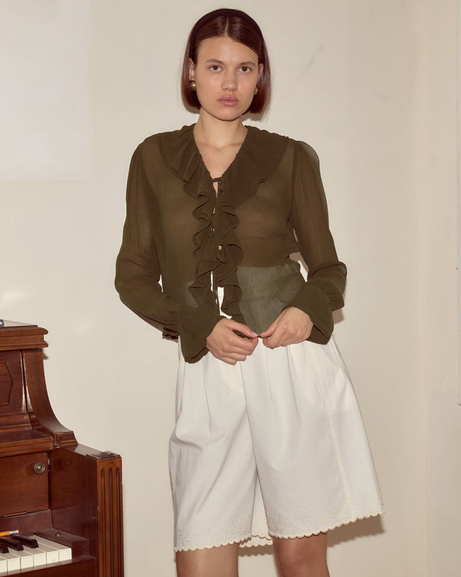 A woman in a Flounce Blouse - Olive and white shorts stands indoors next to a brown upright piano.