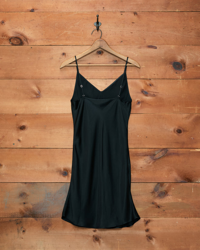 The Lizzie Short Slip - Black, a 100% silk slip dress with spaghetti straps, hangs on a wooden hanger against a wooden wall, evoking the timeless elegance of a vintage 1940s slip.