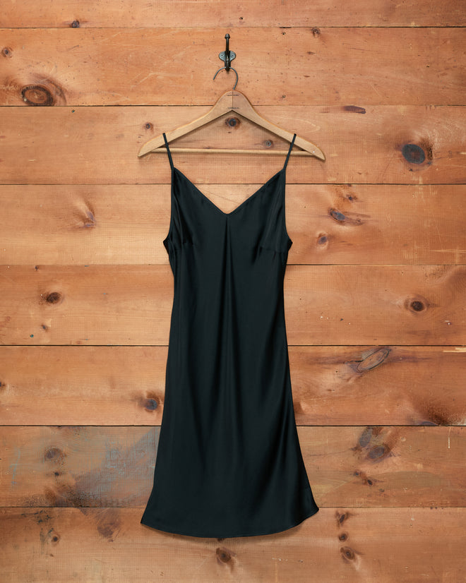 The Lizzie Short Slip - Black, crafted from 100% silk and inspired by 1940s slips, hangs on a wooden hanger against a rustic wooden wall.
