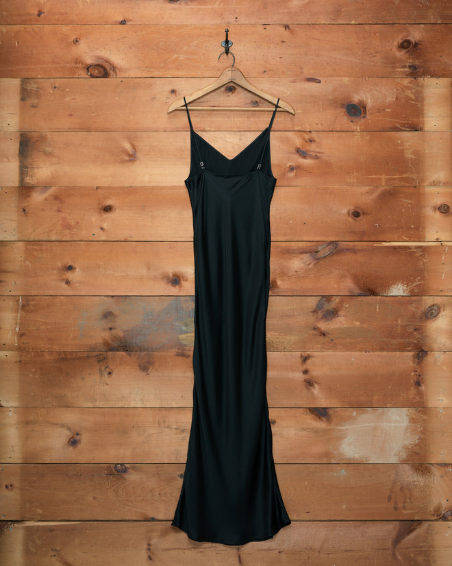 The Lizzie Long Slip - Black, crafted from 100% silk, hangs gracefully on a wooden hanger against a rustic wooden plank wall.