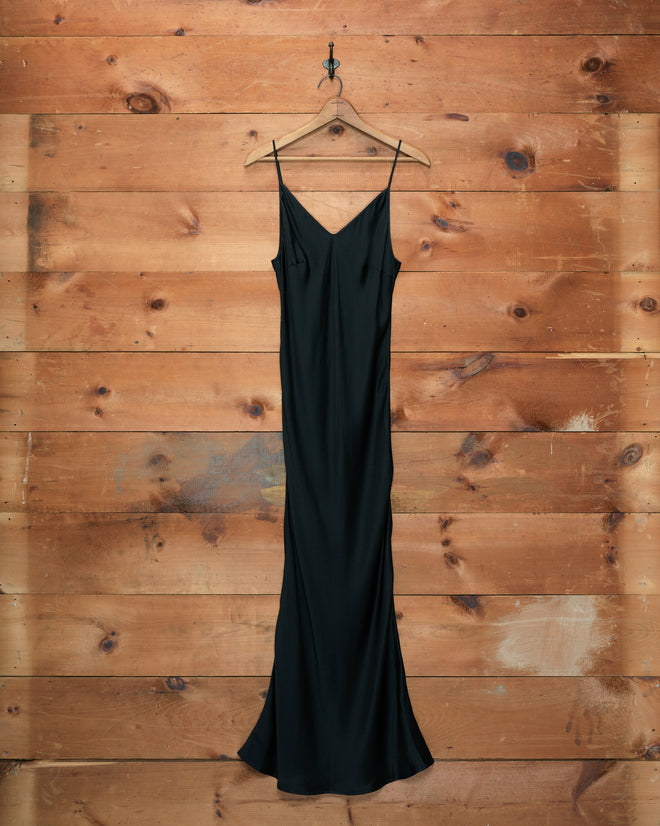 The Lizzie Long Slip - Black, a 100% silk dress inspired by 1940s style, is elegantly displayed on a wooden hanger against a rustic wooden wall background.