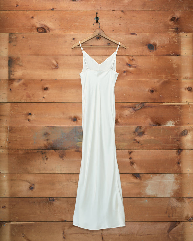 The Lizzie Long Slip - Ivory, crafted from 100% silk, hangs gracefully on a wooden hanger against a backdrop of wooden planks, its vintage-inspired design exuding timeless elegance.