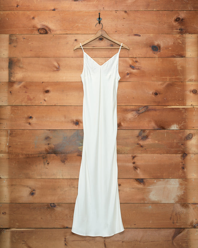 The Lizzie Long Slip - Ivory, a flowing 1940s-style dress crafted from 100% silk, hangs elegantly on a hanger against a wood-paneled wall.