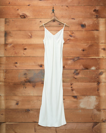 The Lizzie Long Slip - Ivory, a flowing 1940s-style dress crafted from 100% silk, hangs elegantly on a hanger against a wood-paneled wall.