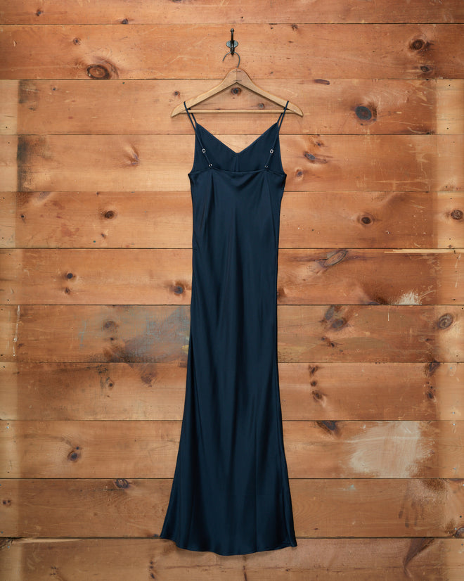 A Lizzie Long Slip in Midnight, crafted from 100% silk, hangs elegantly on a wooden hanger against a wooden wall.