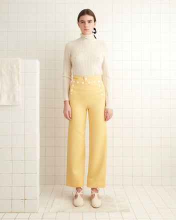 Dressed in a cream sweater and Yellow Riding Twill Sailor Trousers, a person stands in a tiled room, accompanied by white shoes and a towel hanging nearby.