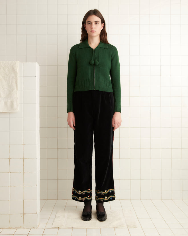 In a tiled room, a person stands wearing a green sweater and the Alpineflower Murray Trousers, distinguished by intricate handmade wildflower embroidery.