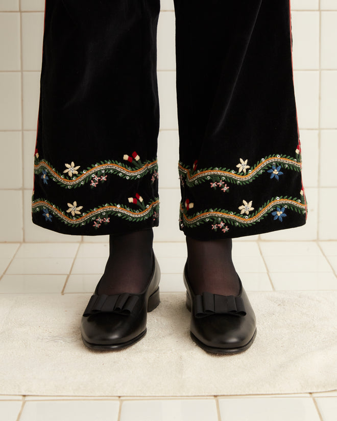 In a tiled room, a person stands wearing a green sweater and the Alpineflower Murray Trousers, distinguished by intricate handmade wildflower embroidery.