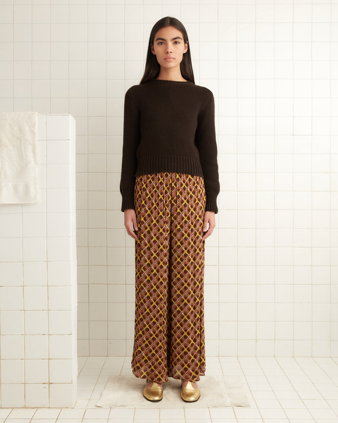 A woman in a dark sweater and Cattail Plaid Pants, inspired by the wide-leg trousers of the 1930s, stands in a tiled room beside a partition.