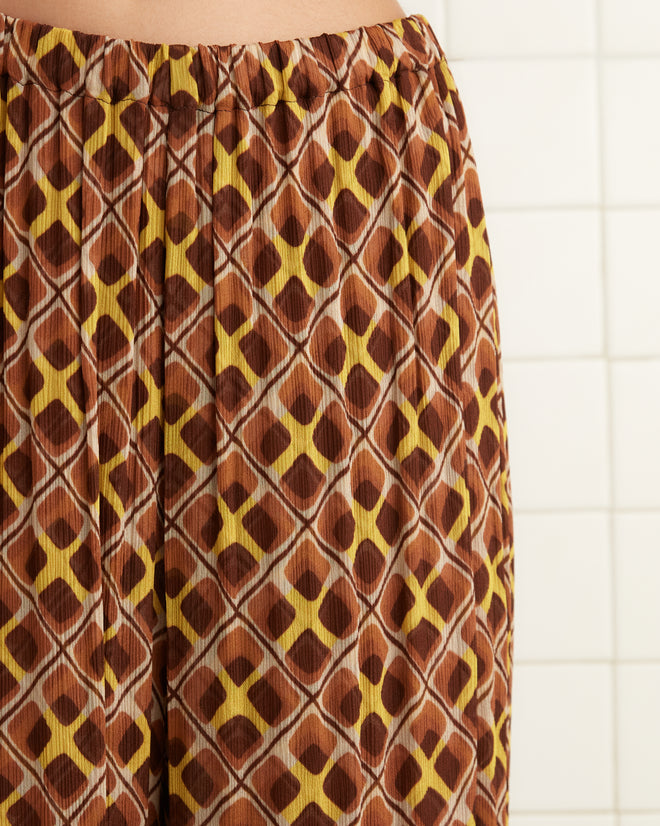 A woman in a dark sweater and Cattail Plaid Pants, inspired by the wide-leg trousers of the 1930s, stands in a tiled room beside a partition.