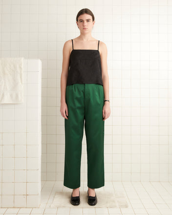 A person stands in a tiled room, facing forward, dressed in a black top and black shoes. They are wearing the Sheen Murray Trousers in green, which feature a satin weave and wide leg design that adds a sophisticated touch to the outfit.