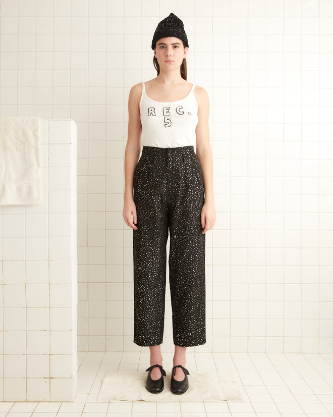 A person is standing in a tiled room wearing a white tank top, the Glitter Murray Trousers, a beanie, and shoes. The trousers are adorned with eye-catching glitter dots that add a touch of sparkle to the look.