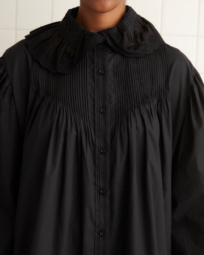 A person in the Petaluma Dress - Black, which features a pintucked bodice, stands in a tiled room wearing knee-high socks with a white towel on a rack.