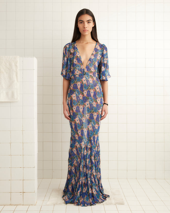 A woman in a 1930s-inspired Daisy Fair Gown, featuring a floral deep V-neck and floor-length design, stands gracefully in a tiled room.