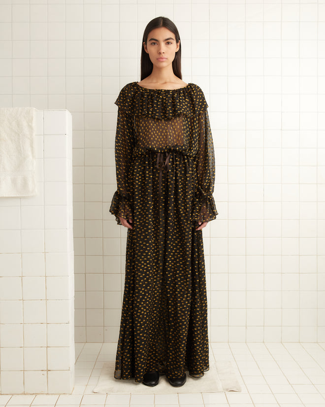 A woman in the Stadium Dot Gown, a long sheer black dress with yellow polka dots that evoke the elegance of a French blouse, stands gracefully in a tiled room.