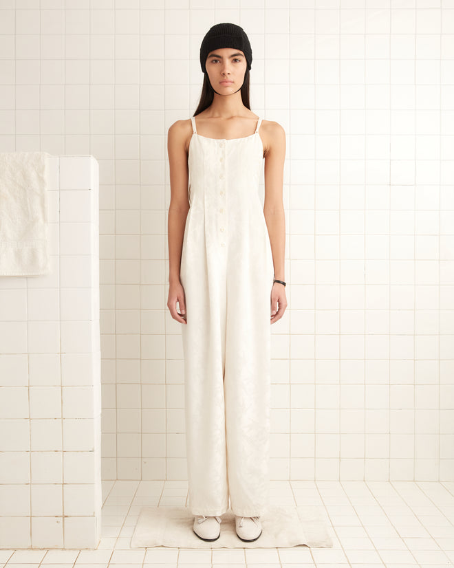 A person in a Football Jacquard Jumpsuit - Cream and a black beanie stands on a tiled floor, facing forward.