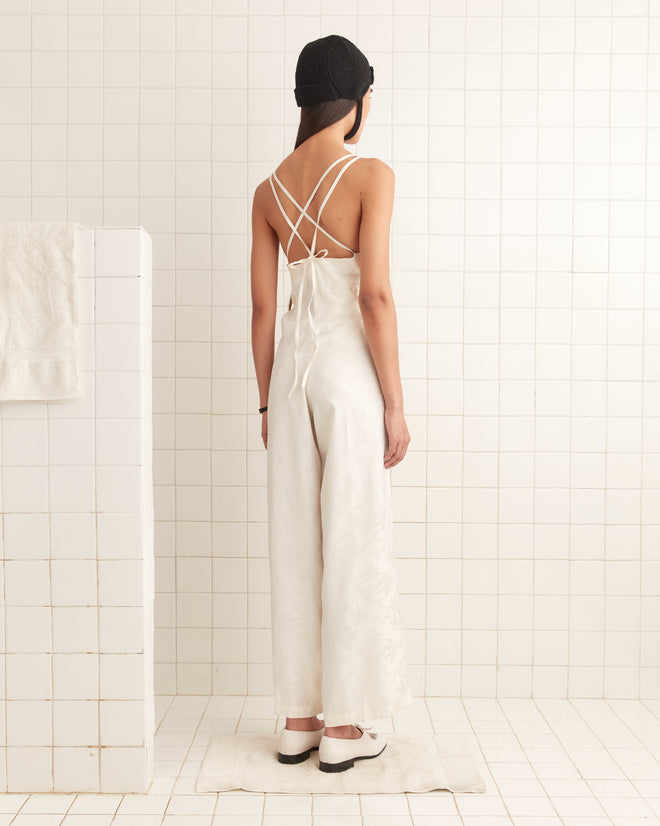A person in a Football Jacquard Jumpsuit - Cream and a black hat stands on a mat in a tiled room, facing away with an air of midcentury elegance.