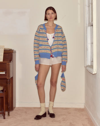 A woman wearing the Wedgewood Cardigan, blue and pink floral virgin wool, with white shorts, socks, and loafers stands by a piano and window.