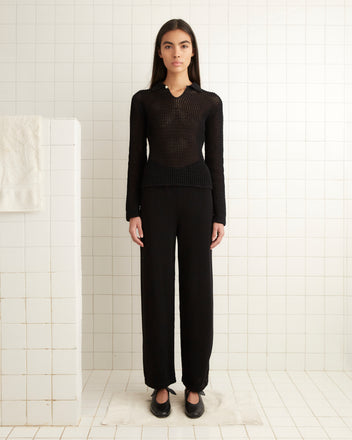 A woman stands in a tiled room wearing a black knit top, Johnny Knit Pants in black, and black shoes.