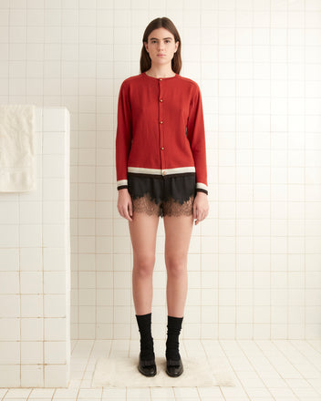 A person in a red Fairground Cardigan and black shorts stands in a tiled room near a white pillar.