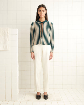 A person with long hair stands in a white-tiled room, elegantly dressed in the Hogan Stripe Cardigan - Green, paired with white pants and black shoes.