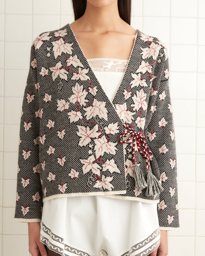 A person stands in a tiled room wearing the Maple Jacquard Cardigan with a floral pattern over a white dress, black tights, and black shoes.