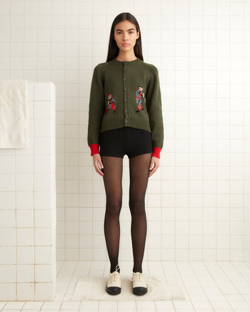 A person stands in a tiled room wearing the Home Run Cardigan, black shorts, sheer tights, and two-tone shoes.