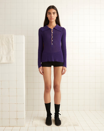 A person wearing the Quincy Top in purple, featuring antique pearl buttons, along with black shorts, stands in a tiled room with a towel on the left.